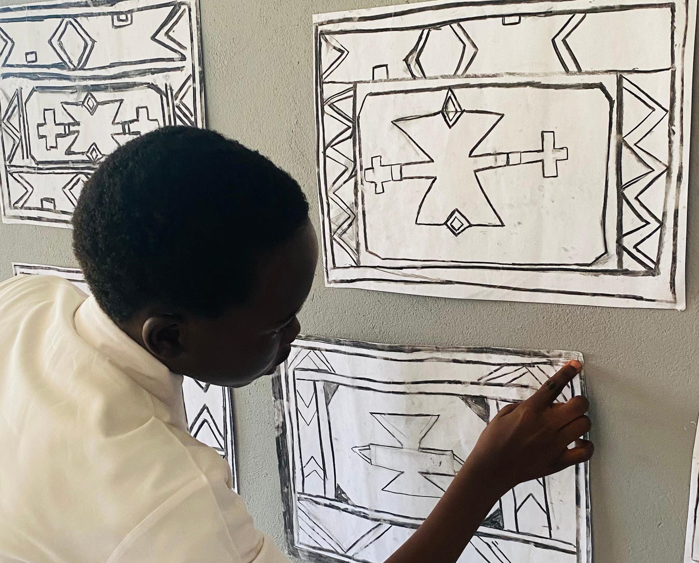 A person in a white shirt attaches a sheet of paper with bold, geometric black-and-white patterns to a wall. Several similar artworks, part of the Legacy Project, are displayed nearby, suggesting a creative or educational celebration.