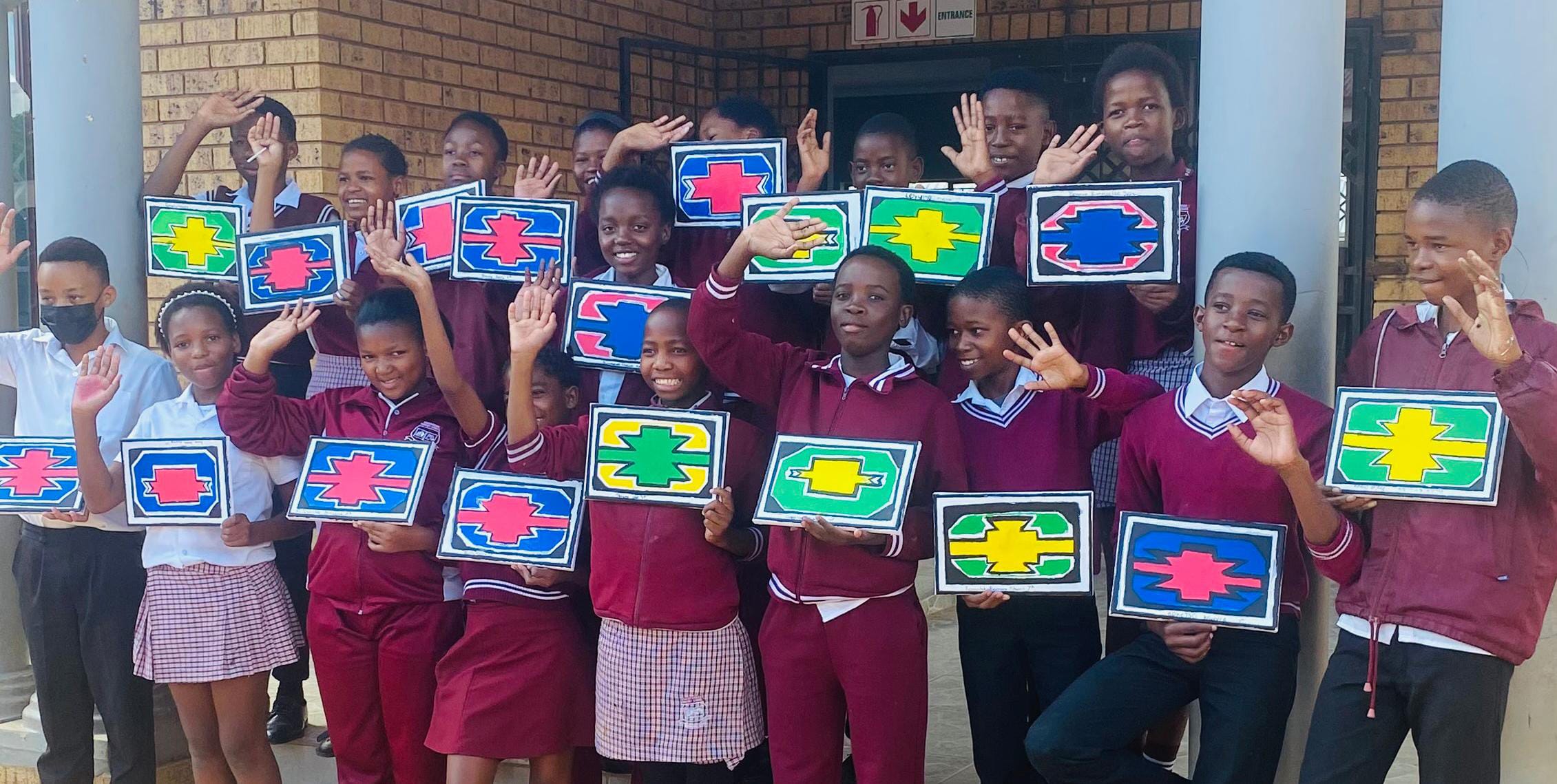 A group of schoolchildren in uniforms stand outside, smiling and waving. Each child holds a colourful rectangular sign, celebrating the Legacy Project with bold patterns. A brick wall is in the background.