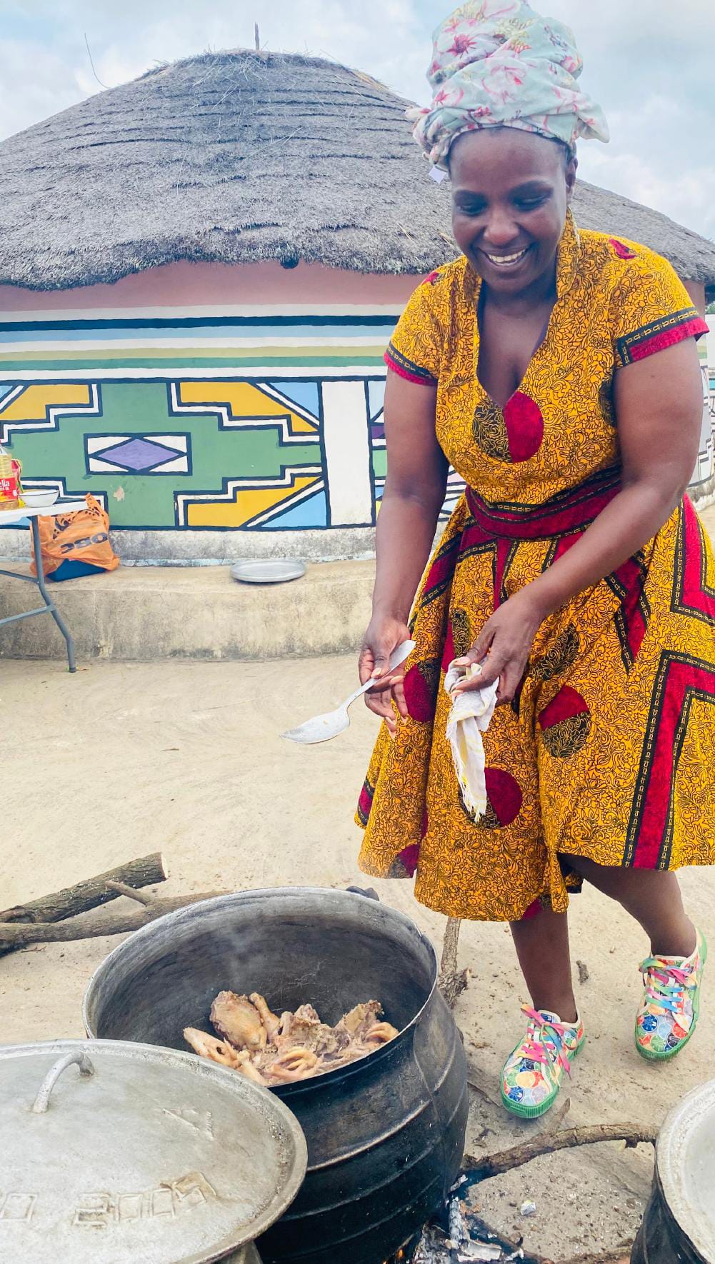Gogo Mahlangu, smiling in a yellow patterned dress and headwrap, cooks meat in a large black pot outdoors. Behind her, a round hut with colourful geometric art sets the scene for this joyful celebration of tradition and community.