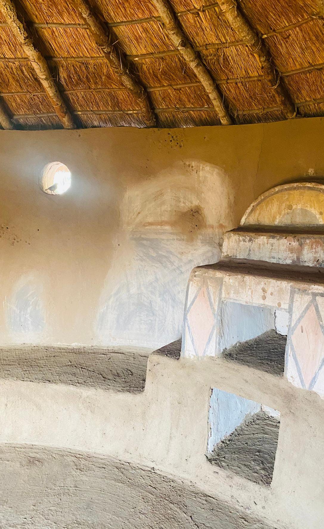 Interior of a traditional mud hut with a thatched roof, inspired by Gogo Mahlangu’s Legacy Project. Earth-toned walls with geometric patterns, natural light from a small window, and built-in mud benches create a rustic, warm atmosphere perfect for celebration.
