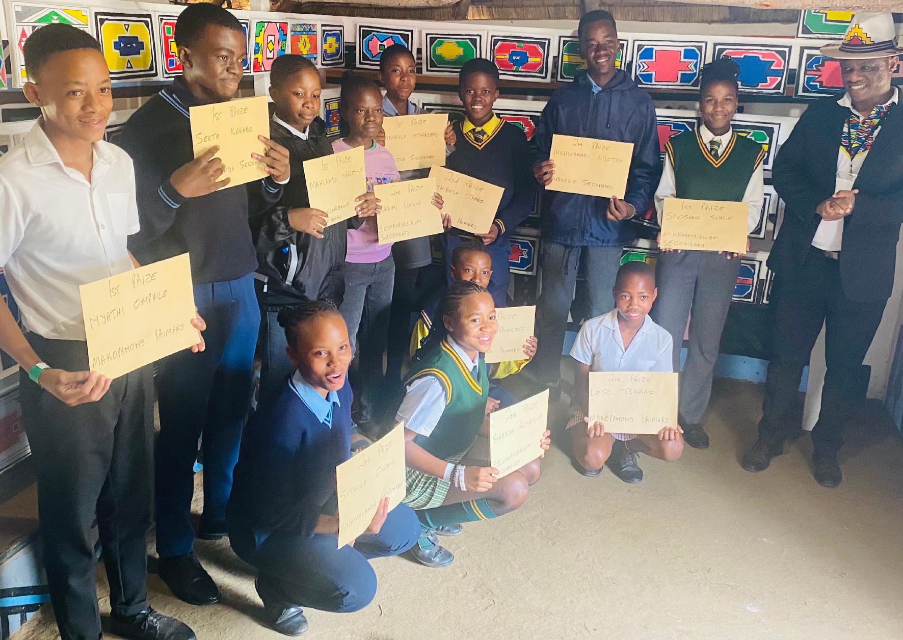 A group of students and two adults pose indoors, smiling and holding certificates during a Legacy Project celebration. The students wear school uniforms, the adults are in formal attire, and colourful artwork adds a vibrant backdrop to the scene.