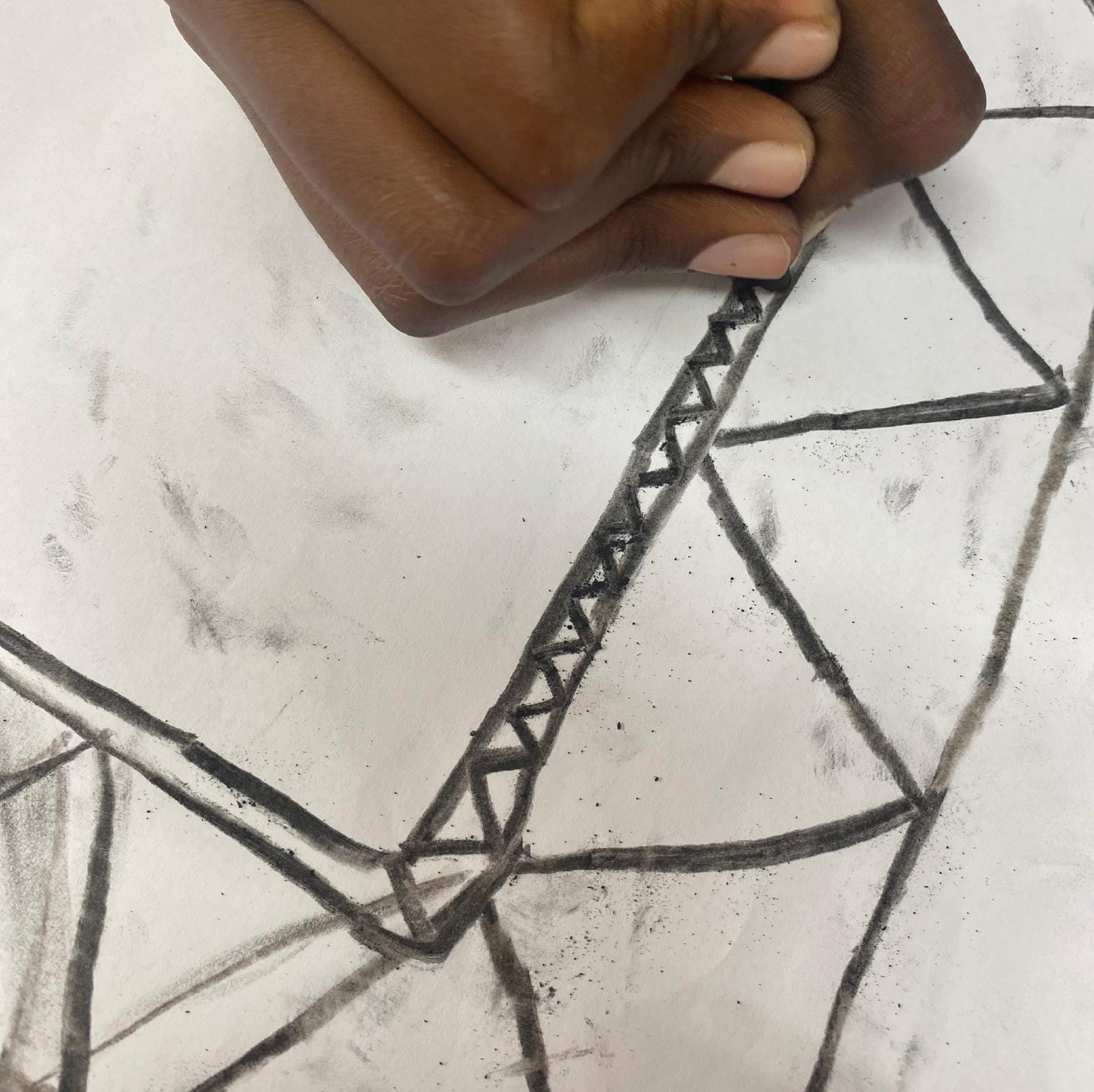 A hand drawing geometric shapes with black charcoal on white paper, reminiscent of a Gogo Mahlangu Legacy Project. The hand adds triangular patterns inside a thick outlined shape; charcoal smudges mark the Celebration of creative expression.
