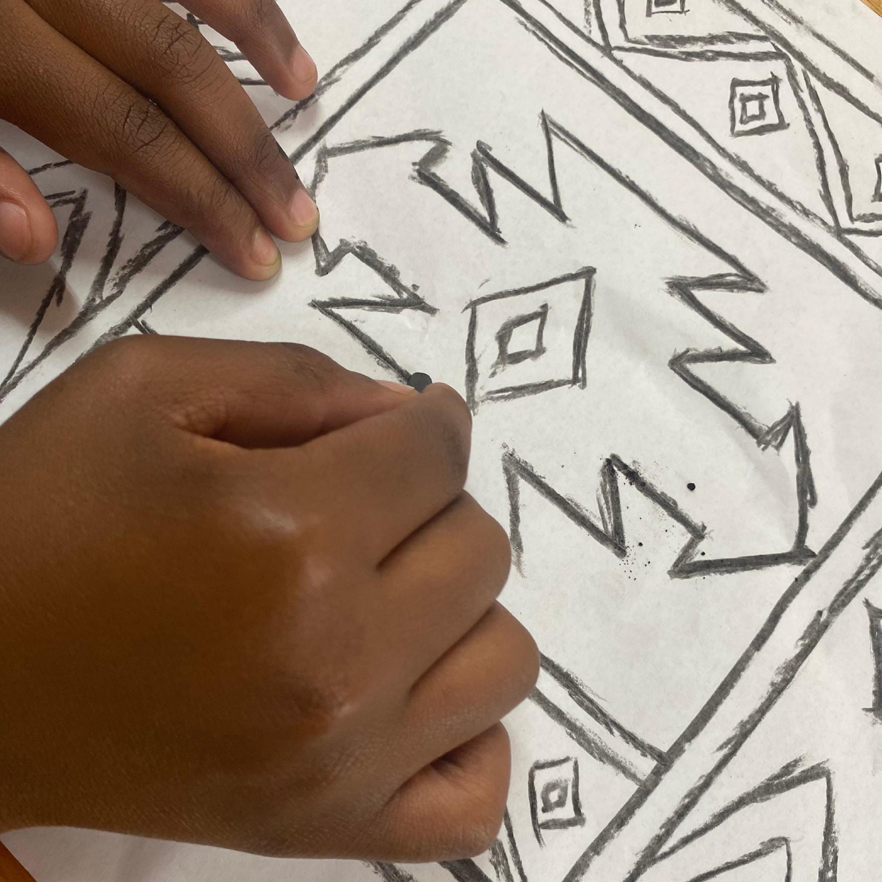 A close-up of two hands drawing bold geometric patterns with a pencil on white paper. Inspired by Gogo Mahlangu, the Legacy Project design features sharp zigzags, triangles, and diamond shapes outlined with thick, dark pencil lines.
