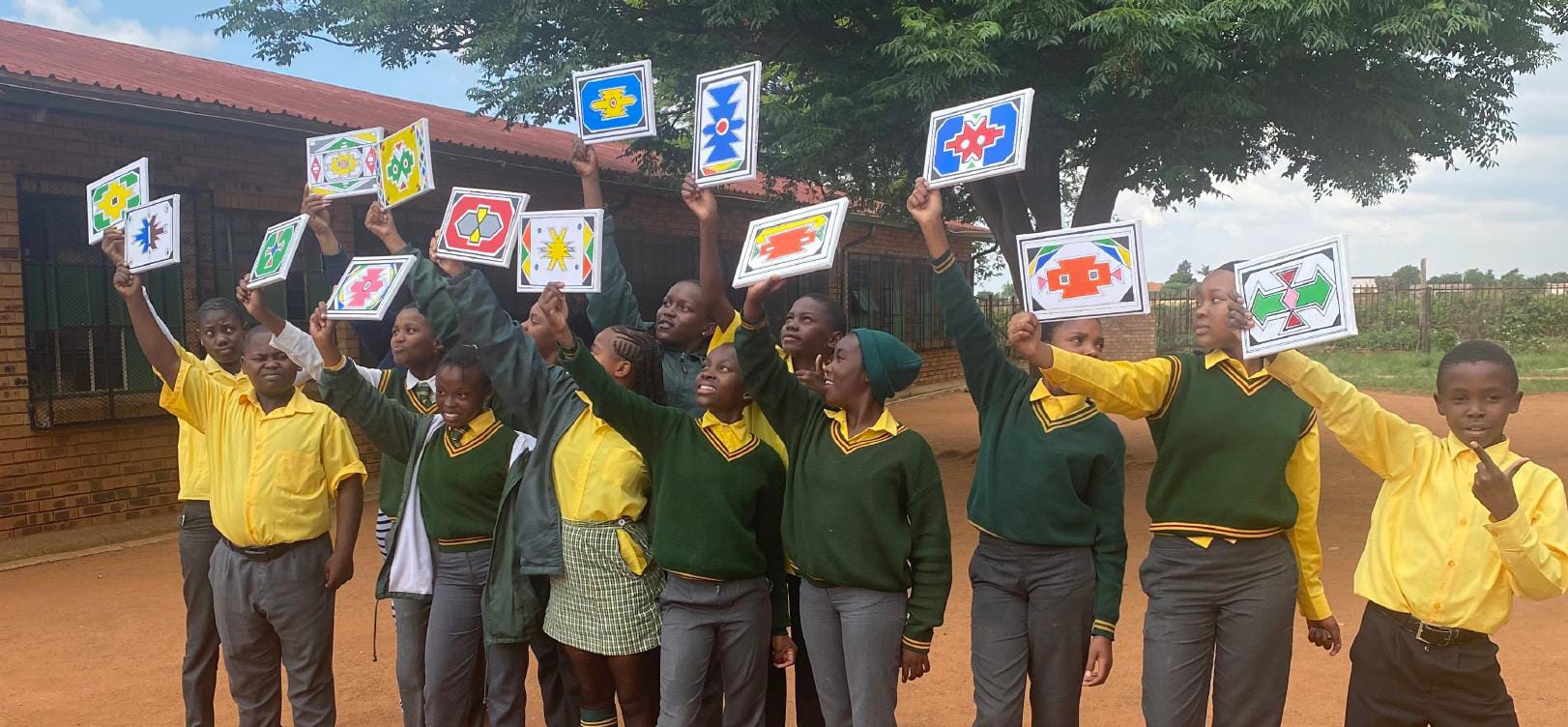 A group of schoolchildren in yellow shirts and green jumpers stand outside, smiling and raising colourful pictures with geometric designs as part of the Legacy Project celebration. They pose in front of a brick building, beneath a large leafy tree.