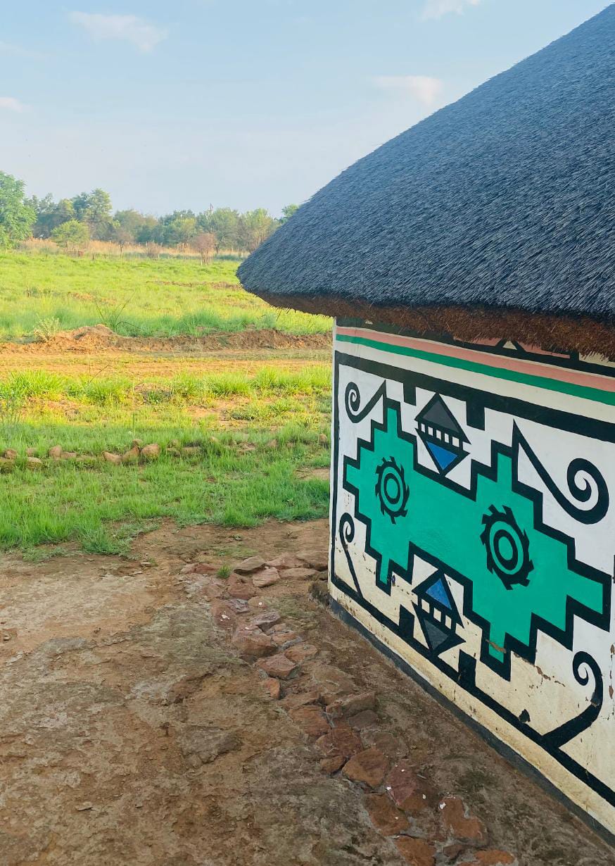 A traditional hut with a thatched roof and a white wall adorned with colourful geometric patterns in green and black celebrates Gogo Mahlangu’s artistry. In the background, lush green fields stretch out under a clear blue sky—a true Legacy Project.