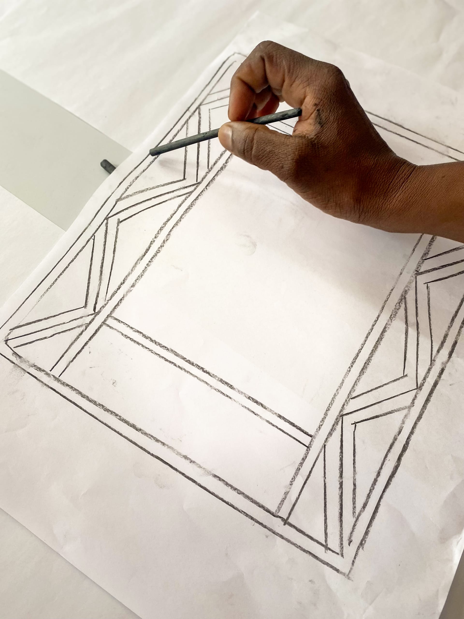 A hand holds a pencil and draws geometric patterns on a white sheet of paper. The design, part of a Gogo Mahlangu Legacy Project, features straight lines and angles forming a decorative border around the paper's edge.