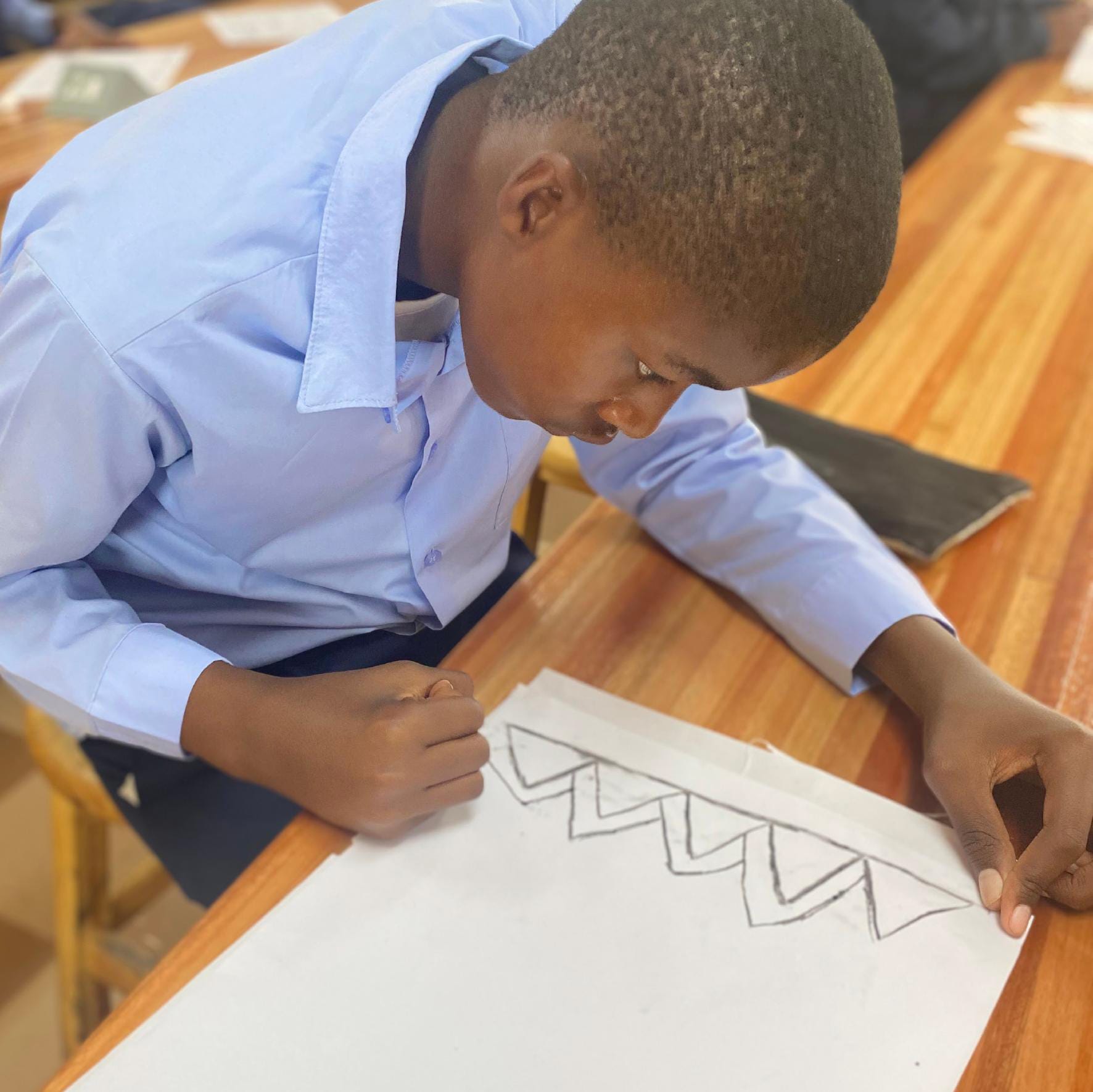 A boy in a light blue shirt sits at a wooden desk, focused on drawing zigzag patterns for the Gogo Mahlangu Legacy Project. His left hand holds the paper steady as he leans in, concentrating on his artwork in a vibrant classroom setting.