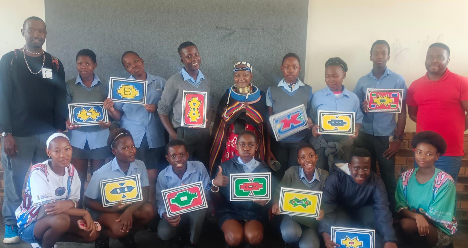 A group of students and adults pose indoors, smiling and holding vibrant geometric artwork for the Legacy Project. Most students wear uniforms; two adults stand at the back, with Gogo Mahlangu in traditional attire centred. A joyful celebration fills the plain classroom background.