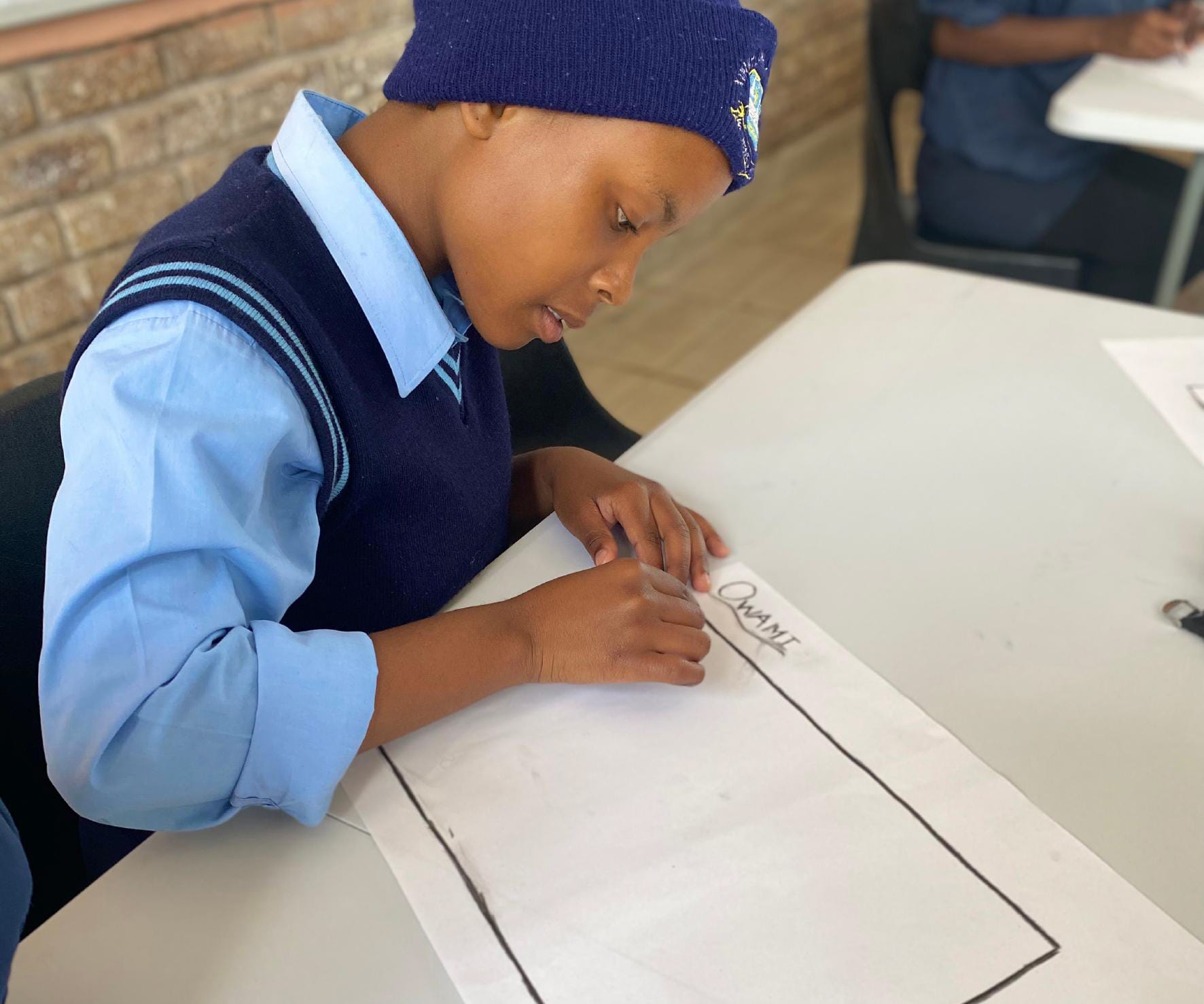 A student in a blue uniform and knitted hat sits at a table, deeply focused as they draw or write on a large sheet of paper labelled 