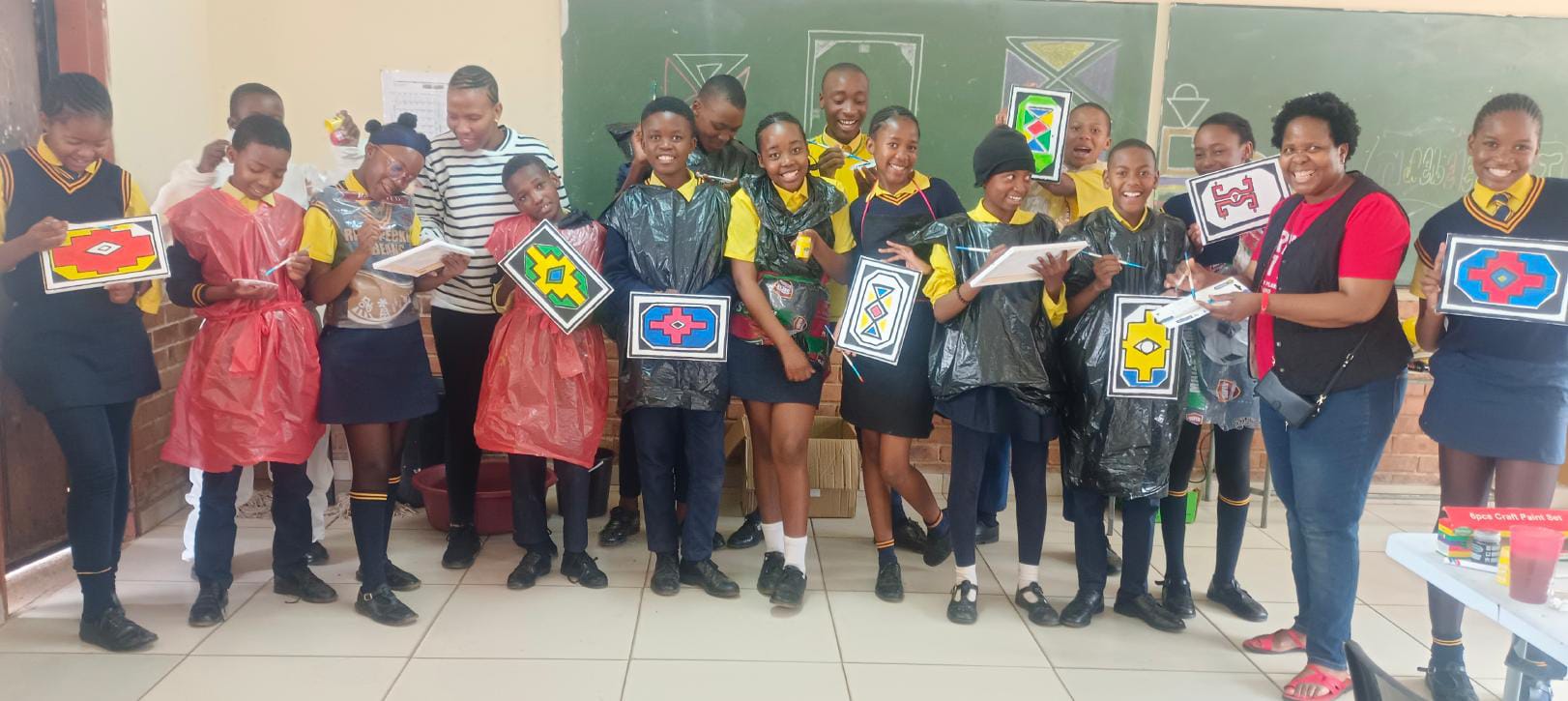 A group of smiling students and a teacher stand indoors, holding colourful geometric artwork. As part of the Legacy Project, some students wear red or black aprons over uniforms. A chalkboard and classroom supplies add to the cheerful, creative classroom scene.