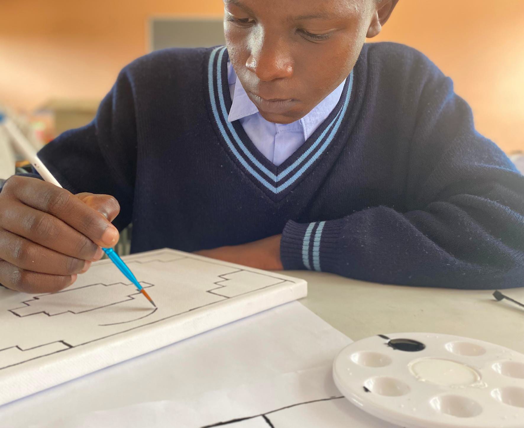A boy in a navy blue school jumper and white shirt paints a geometric design on a white canvas using a blue-handled paintbrush. This creative moment is part of the Gogo Mahlangu Legacy Project, with paint and palette on the table beside him.