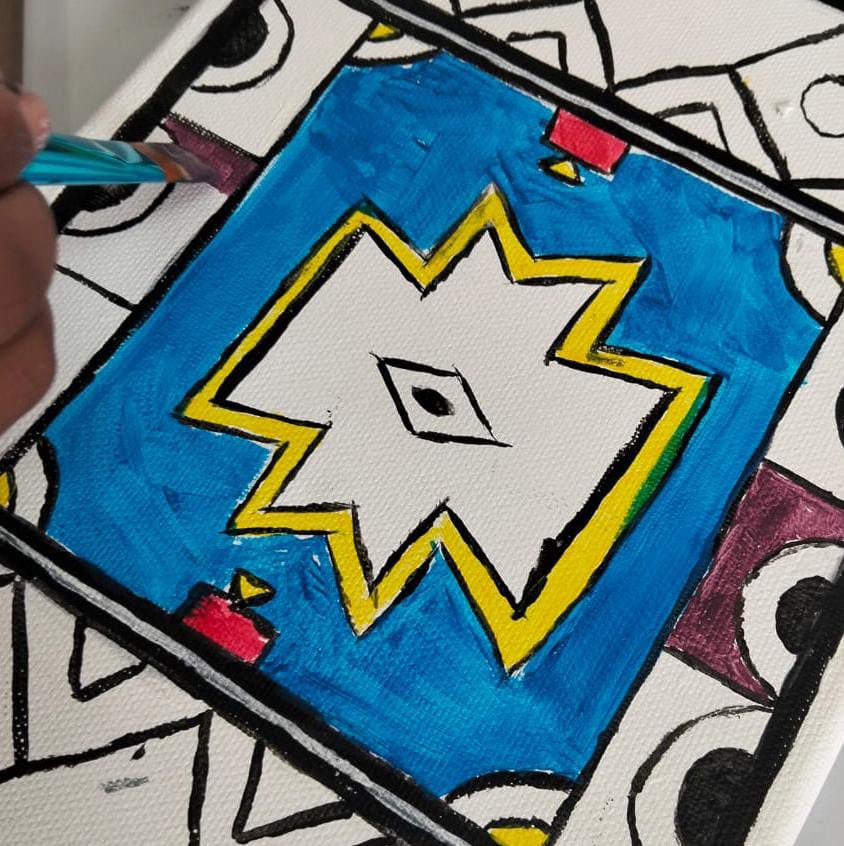 A hand colours a geometric, folk-inspired pattern on canvas in celebration of Gogo Mahlangu. The central motif is a white diamond with a single black eye, outlined in yellow, set against blue and bordered by abstract shapes in red, black, and white.