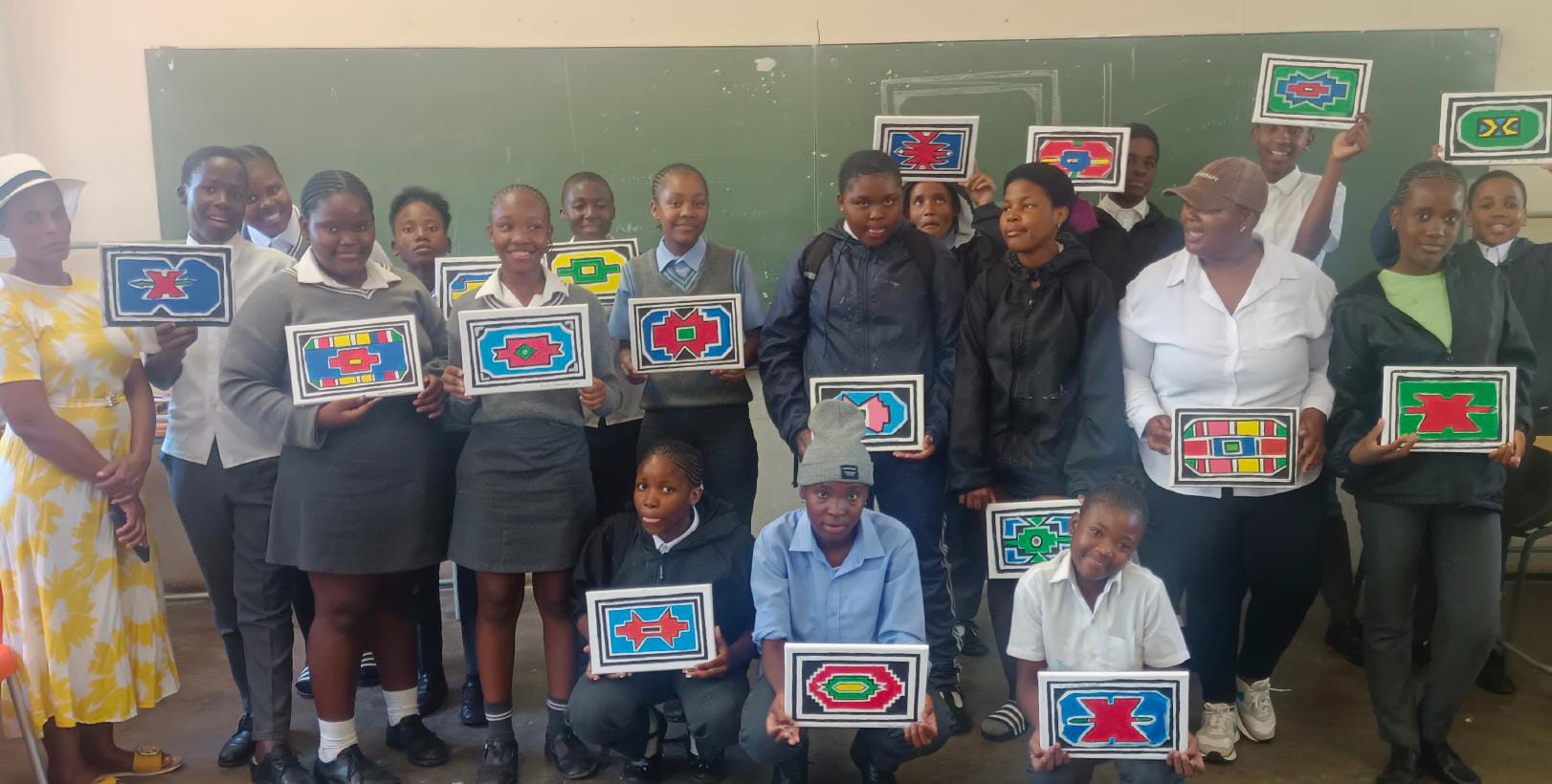 A group of students and two adults stand in a classroom, smiling and holding colourful, rectangular artworks with geometric patterns—a vibrant Celebration for the Legacy Project—posing together in front of a green blackboard.