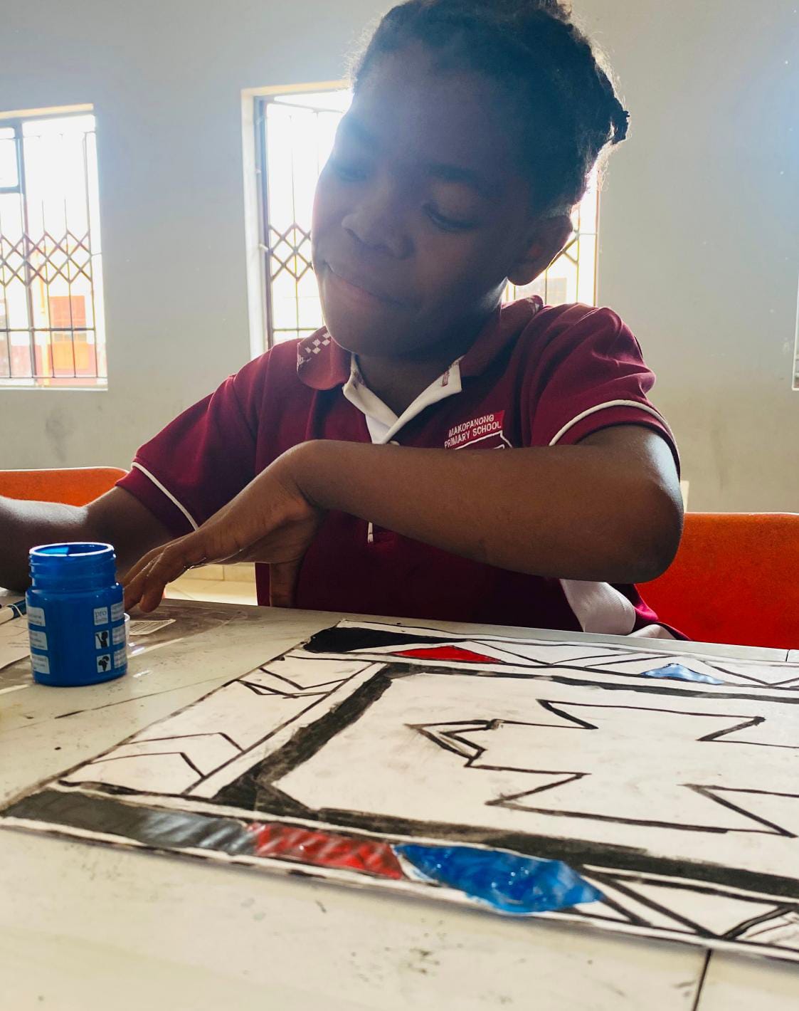 A young girl in a maroon school uniform sits at a table, focused on painting a geometric pattern with black, red, and blue sections—a vibrant scene that captures the spirit of the Legacy Project’s celebration of creativity and bright futures.