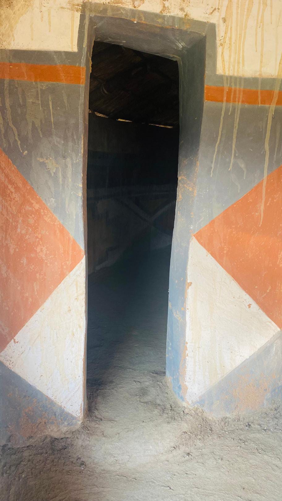 A doorway with thick walls painted in geometric patterns of orange, white, and grey leads into a dimly lit interior. Sunlight highlights the entrance, while faint wall art inside hints at a rich legacy project inspired by Gogo Mahlangu.