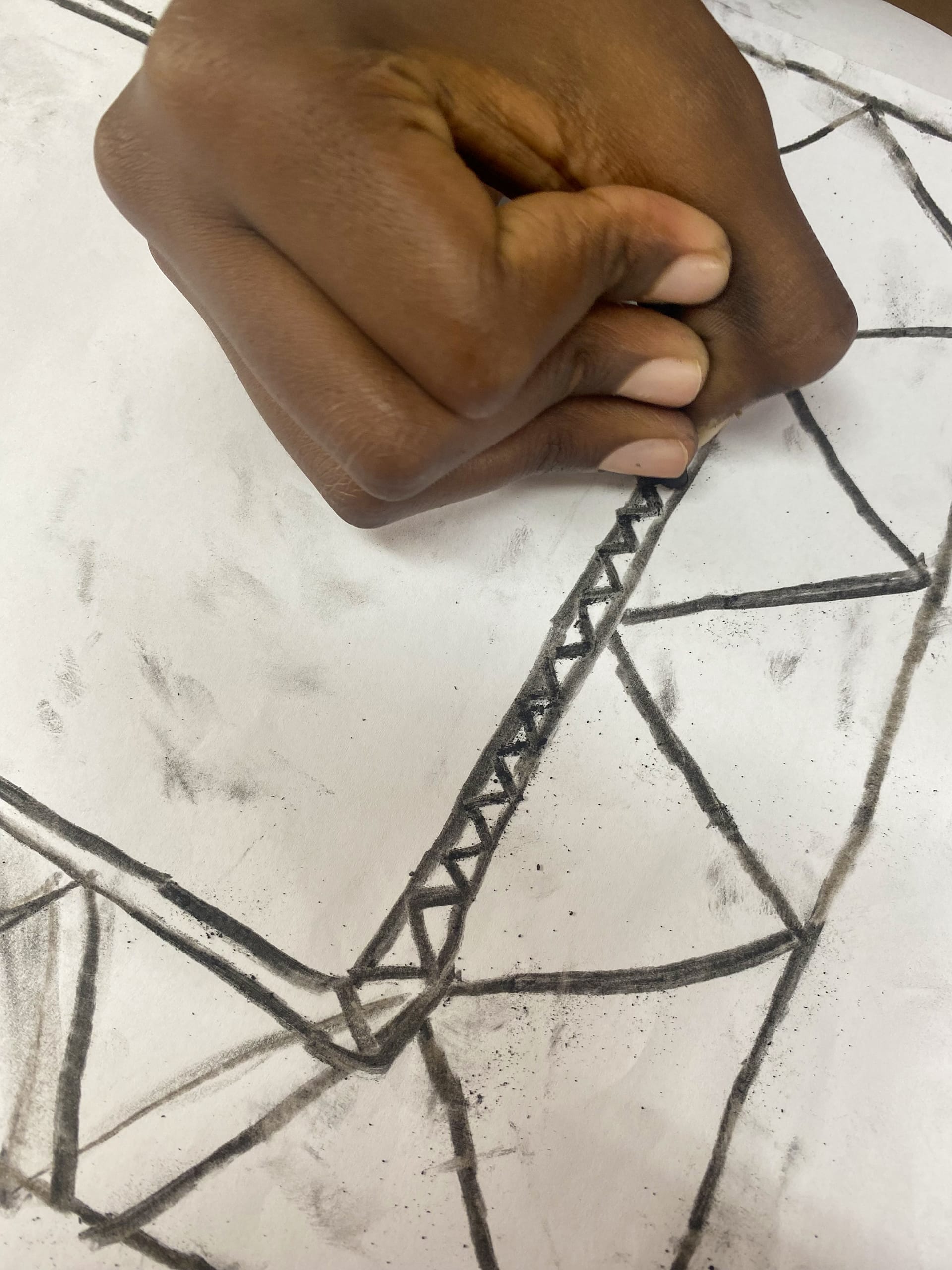 A hand holds a piece of charcoal, drawing a line of small connected triangles within a larger geometric pattern on white paper—part of Gogo Mahlangu’s Legacy Project, where each abstract design becomes a quiet celebration of artistic tradition.