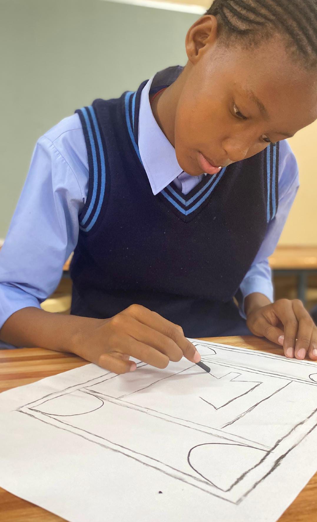 A student in a blue shirt and navy jumper draws geometric shapes, focused on their Legacy Project at a wooden desk. The classroom setting with a blackboard and muted walls adds to the atmosphere of quiet concentration and celebration of learning.