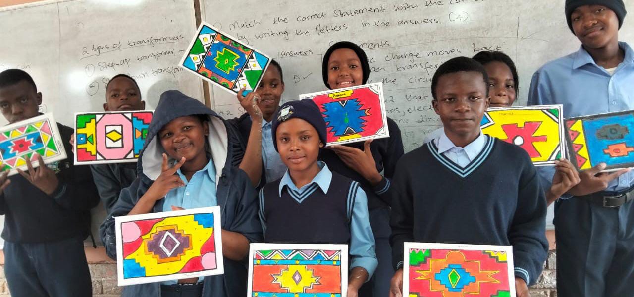 A group of nine pupils in school uniforms stand in a classroom, proudly holding up vibrant geometric artworks created for the Legacy Project. They are smiling at the camera, celebrating their achievements with Gogo Mahlangu’s guidance.