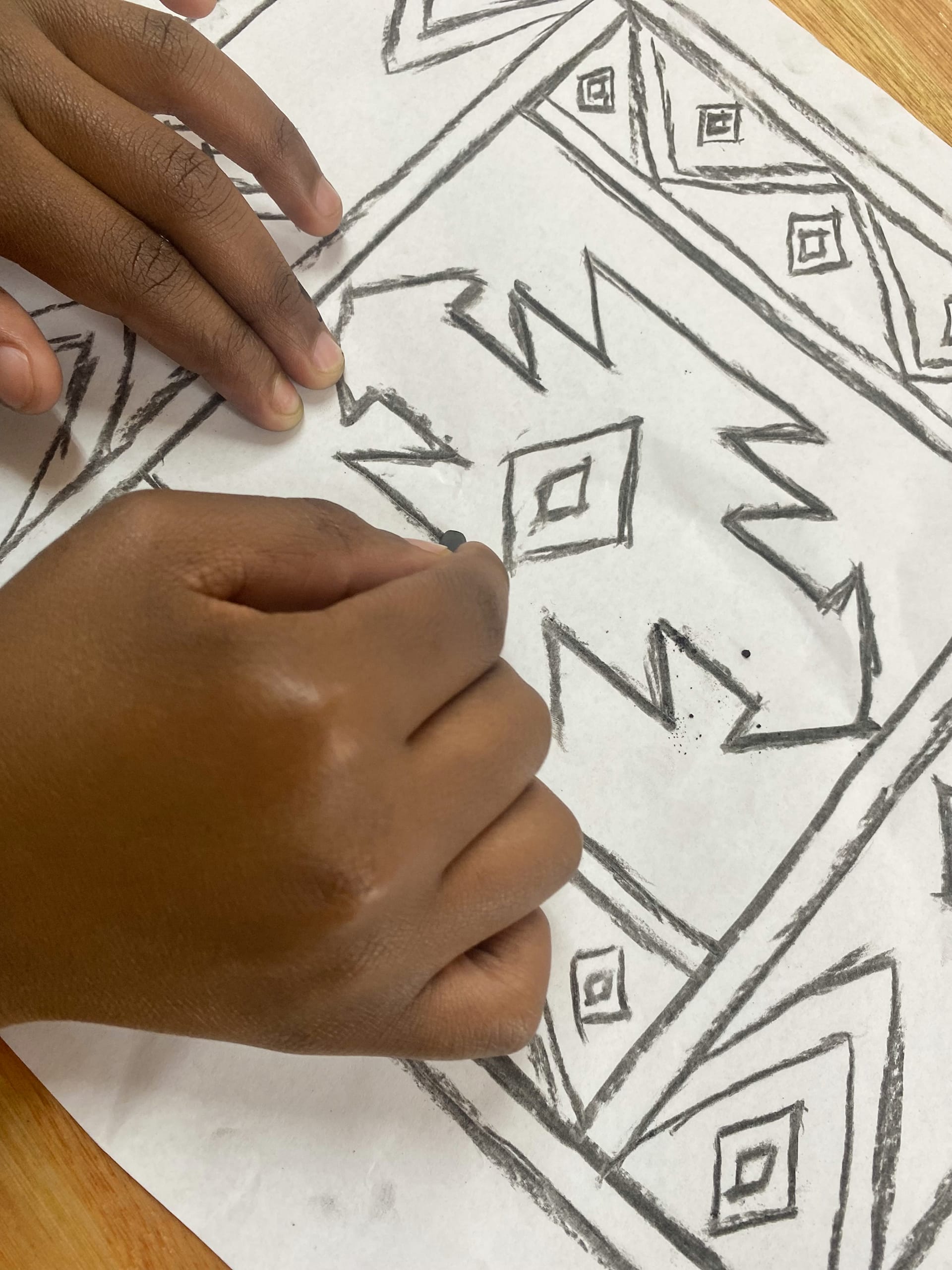 A child’s hands draw bold geometric patterns inspired by Gogo Mahlangu on a white sheet of paper. The Legacy Project design features zigzags, triangles, and diamond shapes. The drawing rests on a wooden surface, with hands at the corners.