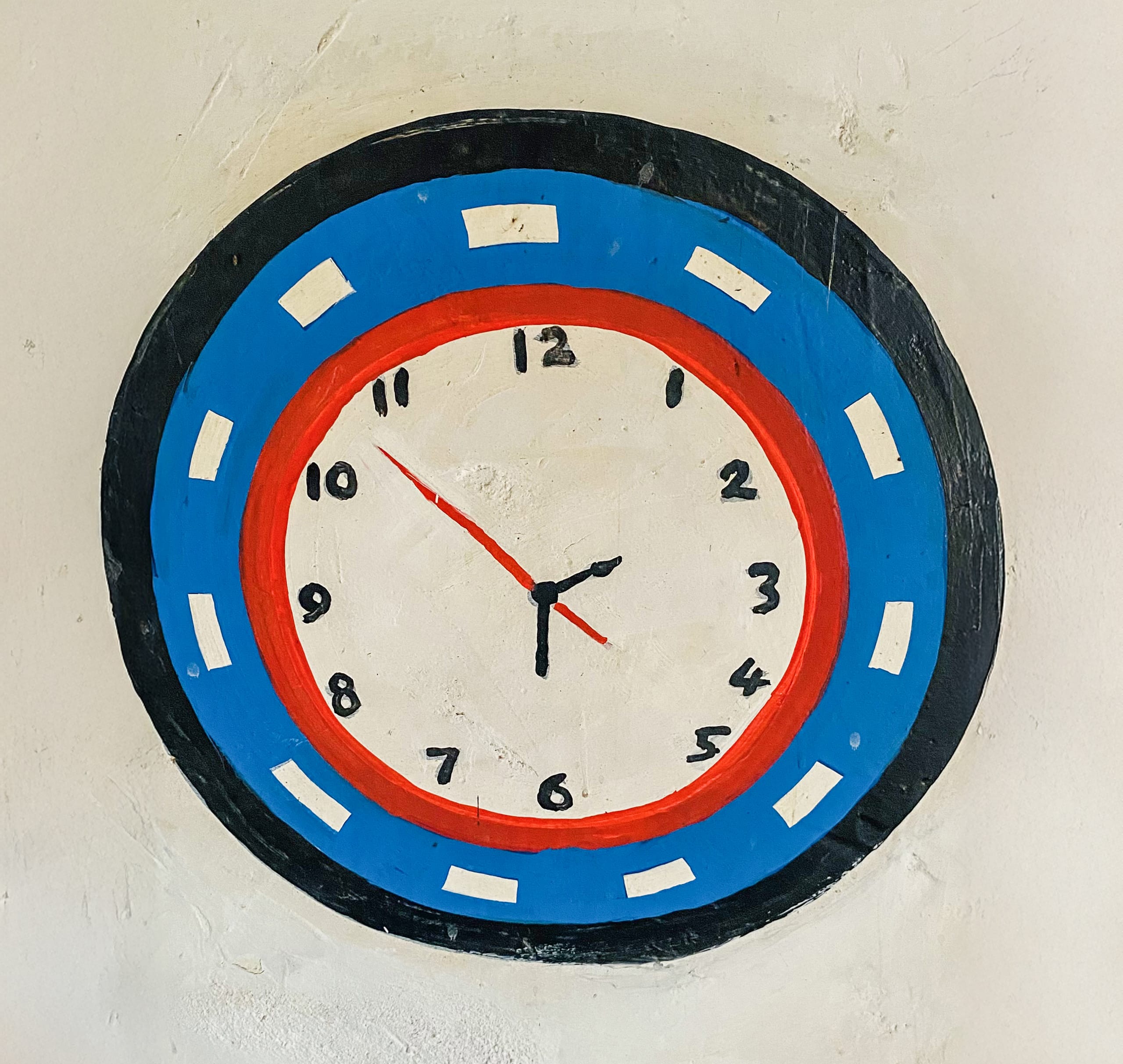 A hand-painted wall clock inspired by Gogo Mahlangu, featuring a black outer rim, blue and red rings, and white face. The design celebrates creativity with simple hour dots and white blocks on the blue ring—a unique Legacy Project piece.