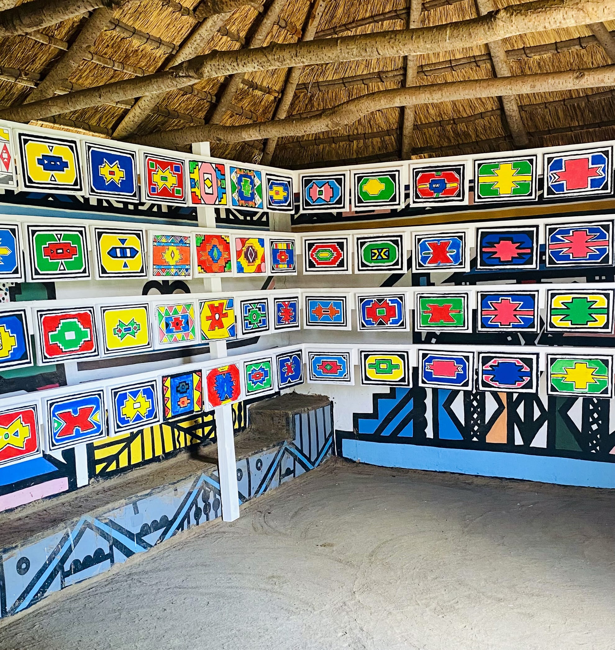 A thatched-roof hut with walls decorated in rows of colourful, geometric Ndebele patterns—a Celebration of Gogo Mahlangu’s artistry. The sandy floor contrasts with the vivid wall art, honouring the Legacy Project’s vibrant heritage.