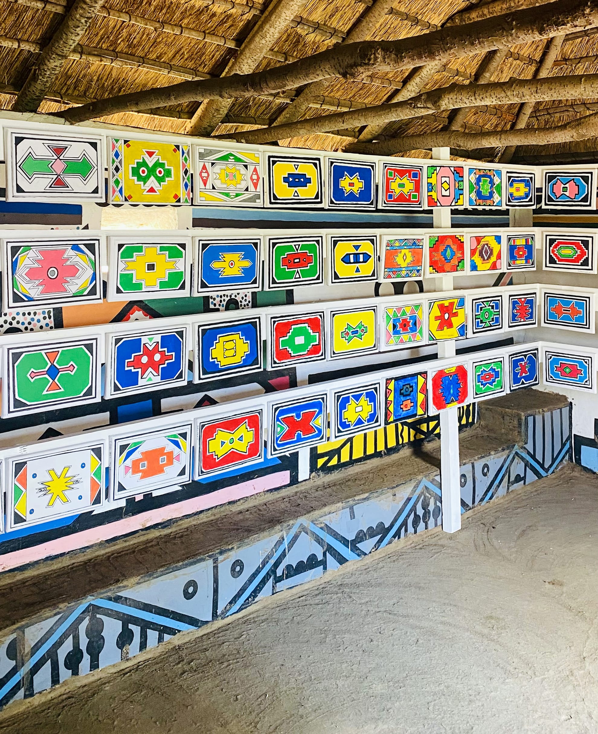 A wall lined with rows of colourful, geometric Ndebele patterns painted on rectangular panels inside a thatched-roof hut—a vibrant celebration of tradition, featuring Gogo Mahlangu’s Legacy Project. The sandy floor and lower black-and-white walls complete the scene.