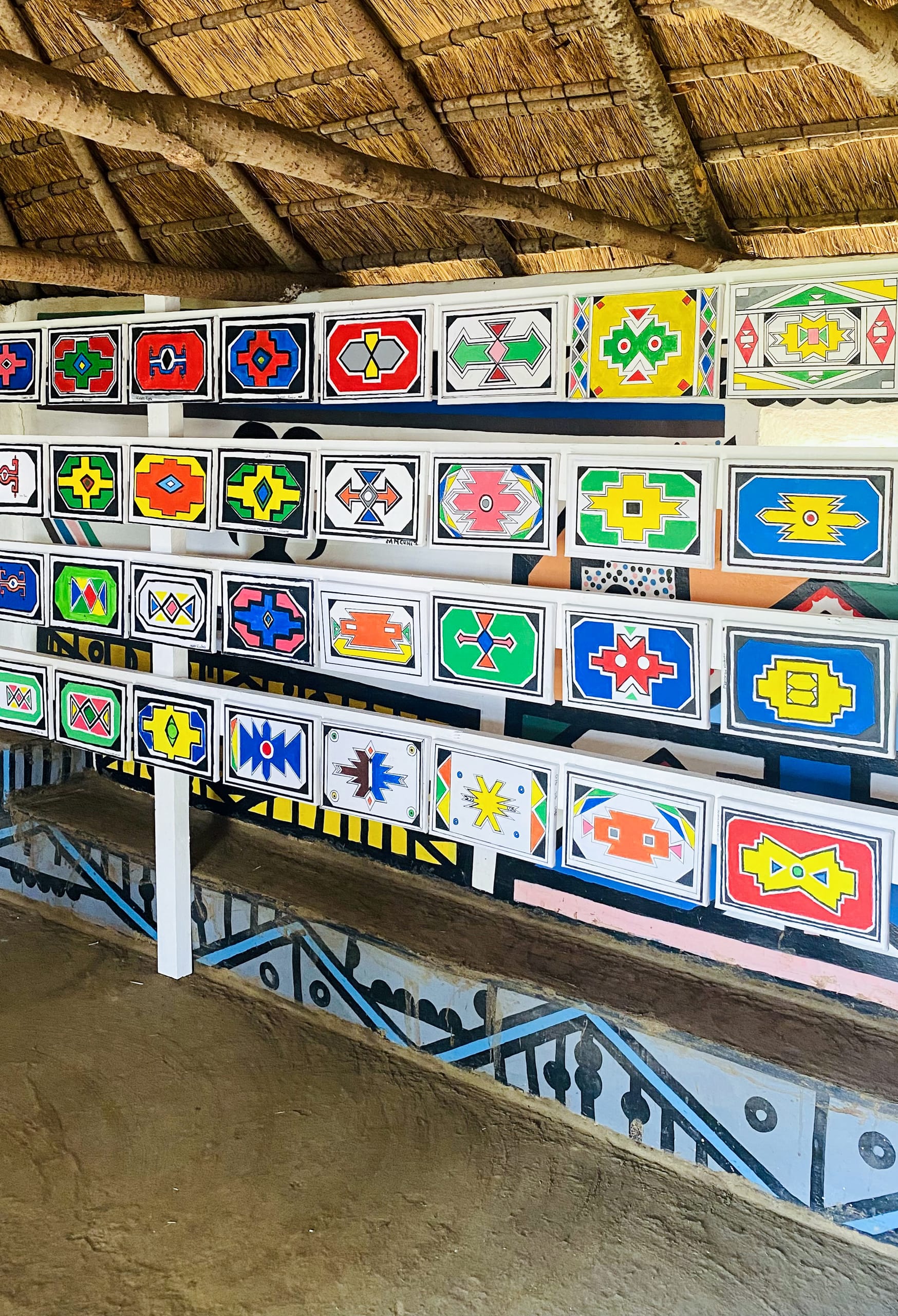 A wall lined with colourful rectangular panels painted in vibrant geometric Ndebele patterns, part of the Legacy Project celebrating Gogo Mahlangu. The floor is brown earth, and the lower wall features striking geometric designs in black, white, and blue.
