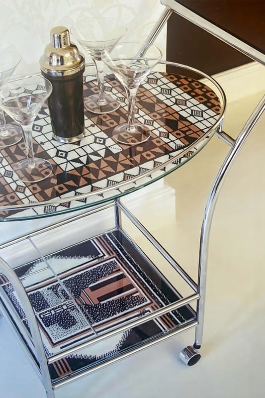 A metal drinks trolley with a geometric patterned glass top holds four martini glasses and a cocktail shaker. Below, the lower shelf displays a similarly patterned tray. The trolley sits on a pale floor near a white wall and skirting board.