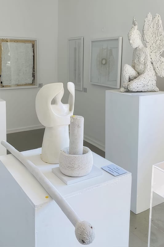 A contemporary art gallery displays abstract white sculptures on plinths, including a smooth, curved bust, a seated winged figure, a rod with a textured ball, and bowl-like forms. Minimalist framed artworks hang on the white walls in the background.