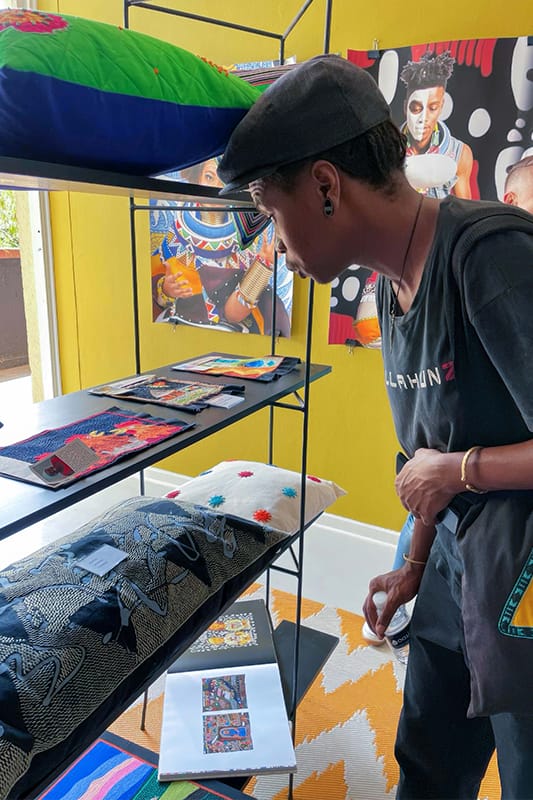 A person in a cap and black shirt leans forward to closely examine textiles and artwork displayed on shelves. Brightly coloured cushions and art pieces fill the shelves; vibrant paintings are visible on a yellow wall in the background.