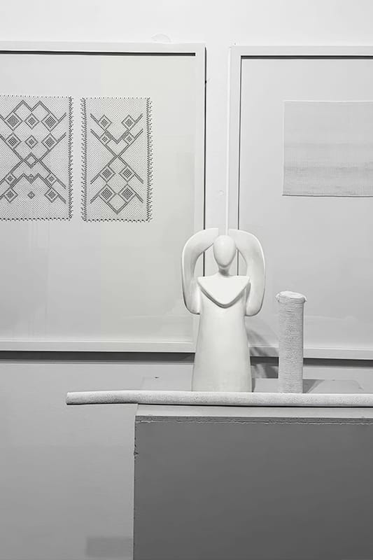 A minimalistic white sculpture of a stylised human figure with raised arms stands on a ledge. Behind it are two framed, geometric textile artworks and a plain fabric piece, all in black, white, and grey tones, creating a modern, abstract display.