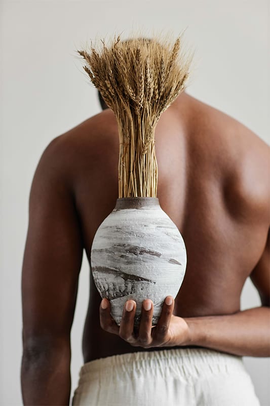A shirtless person stands with their back to the camera, holding a round, textured ceramic vase filled with wheat stalks behind them. The background is neutral and minimal, emphasising the vase and wheat. The person wears light-coloured trousers.