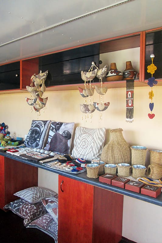 A display of handmade crafts on a wooden counter, including woven baskets, pottery, beaded jewellery, fabric cushions with artistic prints, small boxes, and colourful hanging ornaments. Shelves above hold more crafts and decorative items in a well-lit room.