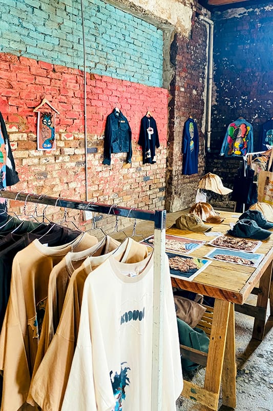 A rustic shop interior with exposed brick walls displays colourful art, T-shirts, and jackets. Clothing hangs on rails and wooden tables, with art prints spread out. Bright shirts and framed art decorate the textured brick wall in the background.