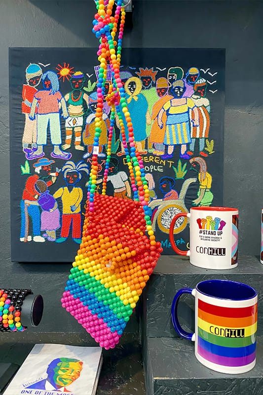 A vibrant display features a beaded rainbow handbag, colourful mugs (one with a rainbow and 