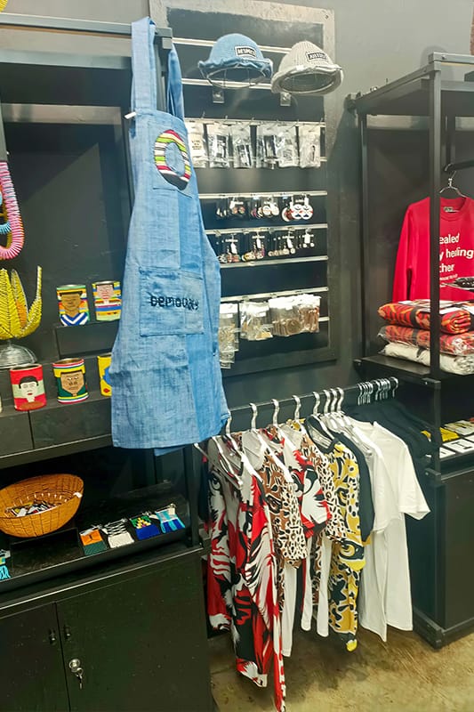 A shop display features colourful shirts, a blue apron with “DEPOP” text, hats, socks, accessories, mugs, a woven basket, and a red sweatshirt folded on a shelf. The background is dark, making the vibrant products stand out.