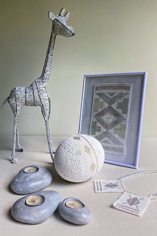 A beaded giraffe figurine, a framed geometric beadwork, a white beaded egg, three smooth stone tealight holders, and two beaded necklaces are arranged on a neutral surface against a light background.
