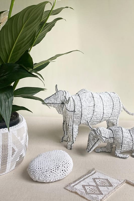 A potted green plant sits next to two beaded animal sculptures, resembling a rhino and a calf, on a beige surface. In front are a round, white woven object and a flat rectangular beaded piece with geometric patterns. The background is light and neutral.