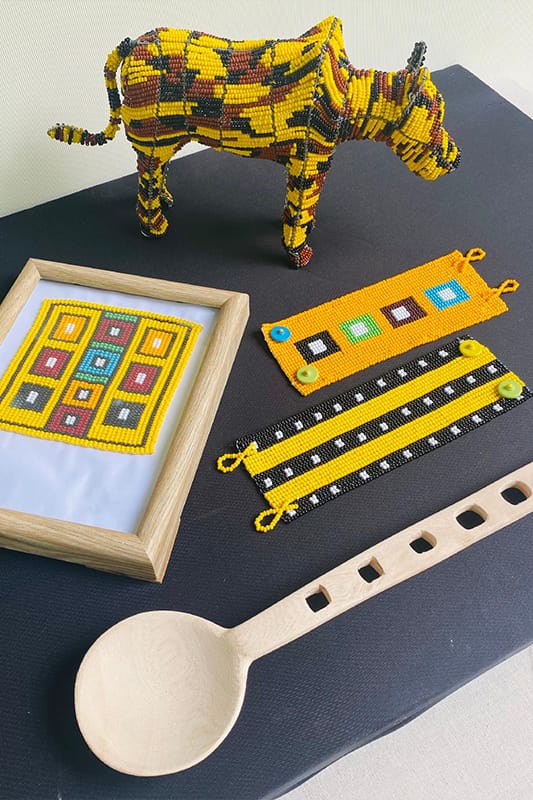 A beaded cow sculpture, a framed geometric beadwork, three colourful beaded bracelets, and a large wooden spoon with square holes are displayed on a black surface against a light background.