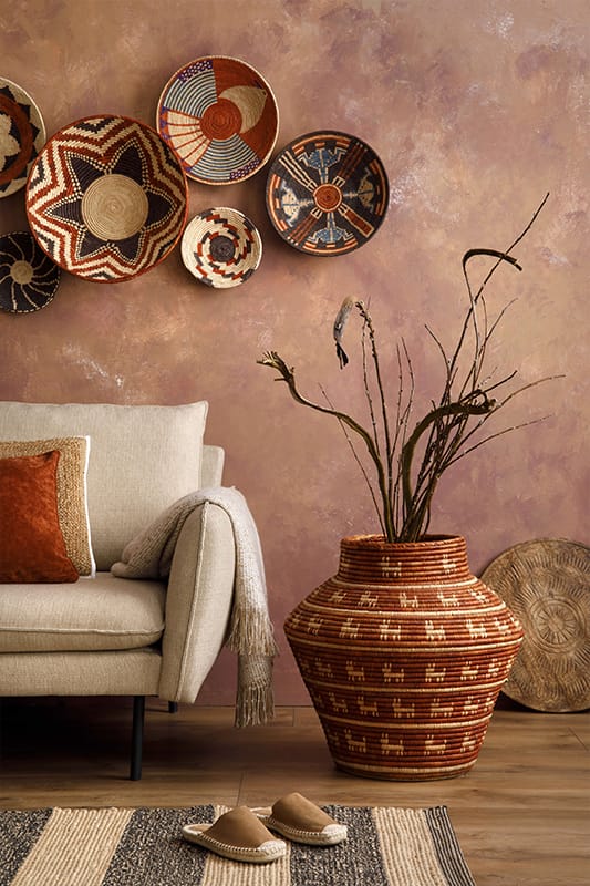 A cosy living room features a beige sofa with brown and beige cushions and a draped grey throw. Above the sofa, five woven baskets with geometric patterns hang on a textured mauve wall. A large decorative basket holds dried branches. Slippers and a rug are on the wooden floor.