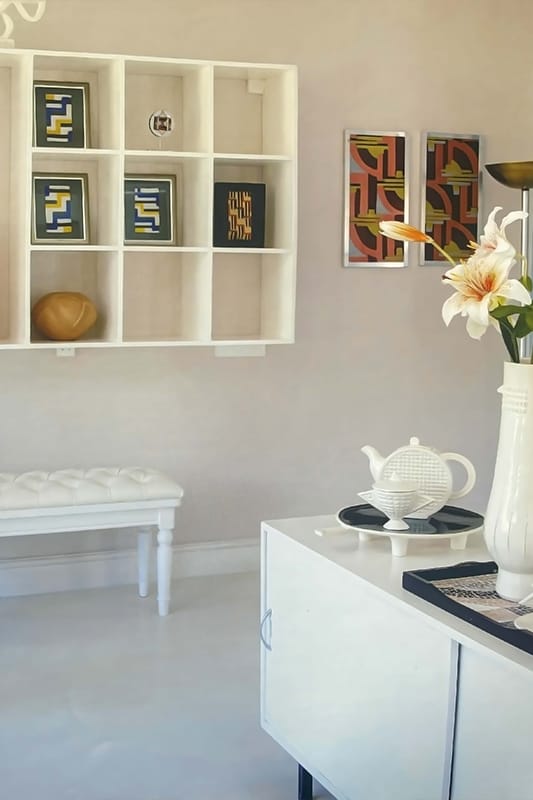 A minimalist room with a white wall shelf holding abstract art and a decorative bowl, a white bench below, and a white cupboard nearby. On the cupboard are a white teapot, a chequered tray, and a vase with lilies. Two colourful abstract paintings hang on the wall.