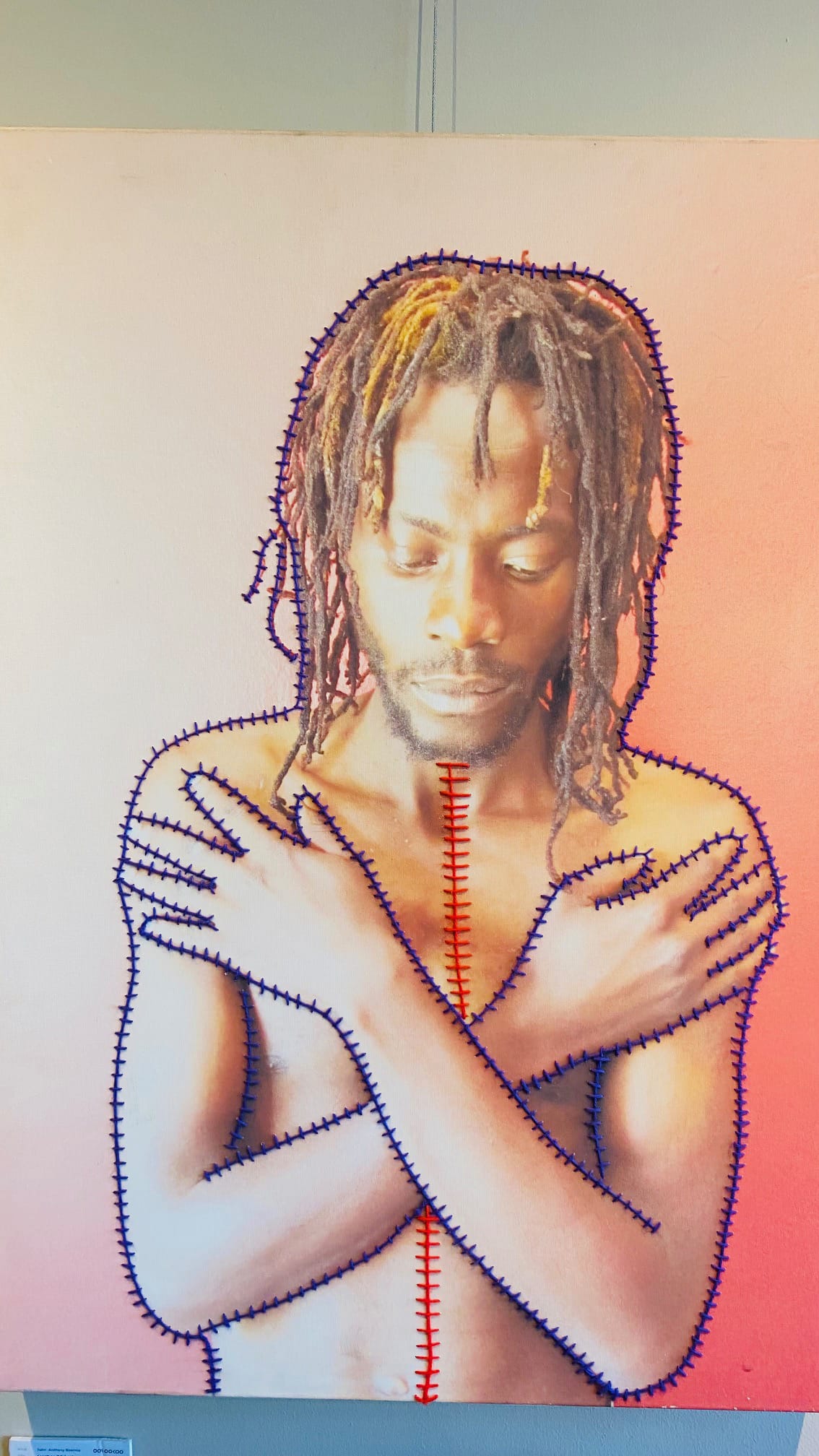 A shirtless person with medium brown skin and dreadlocks stands against a pink background, arms crossed over their chest. Blue and red stitches, drawn or sewn, outline their body and trace a vertical line down the centre and across the arms.