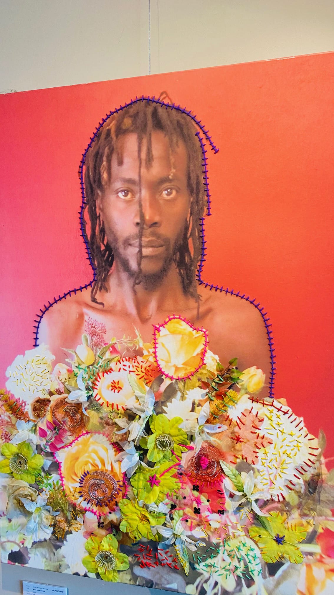 A portrait of a person with medium-length dreadlocks and a neutral expression is set against a bright red background. Colourful, textured flowers and stitched accents surround their shoulders and chest, blending photographic and crafted elements.