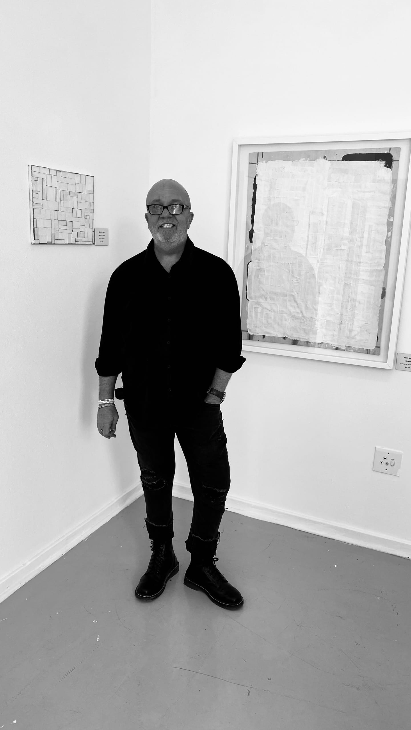 A man with glasses, a beard, and dressed in dark clothing stands in a white-walled art gallery. He is positioned between two abstract framed artworks, one small and one large. The floor is plain, and the space appears minimalistic and modern.