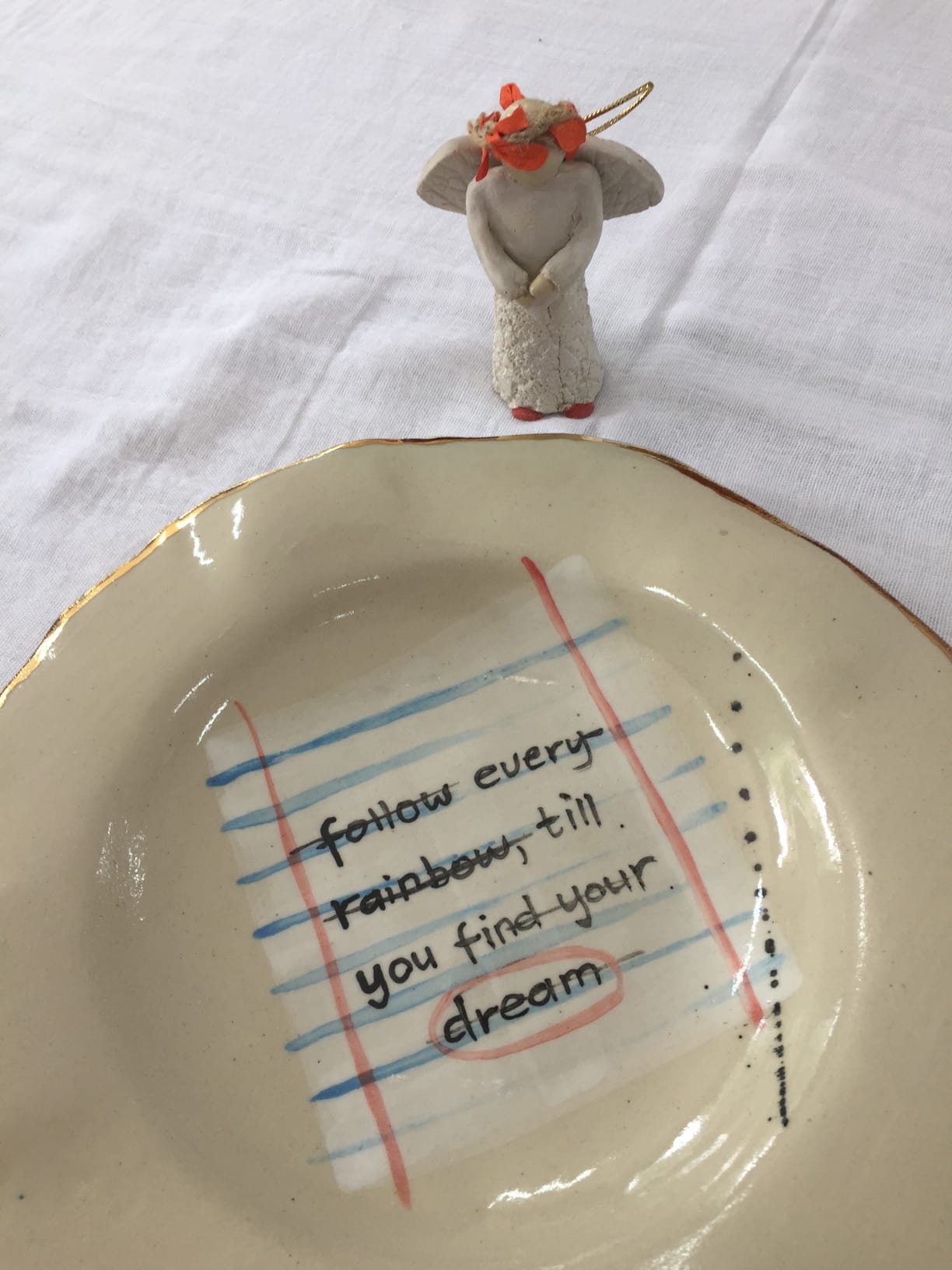 A ceramic plate with a notebook paper design and the words “follow every rainbow, till you find your dream” is shown on a white cloth. At the top, there is a small angel figurine with a red flower crown and hands together in prayer.