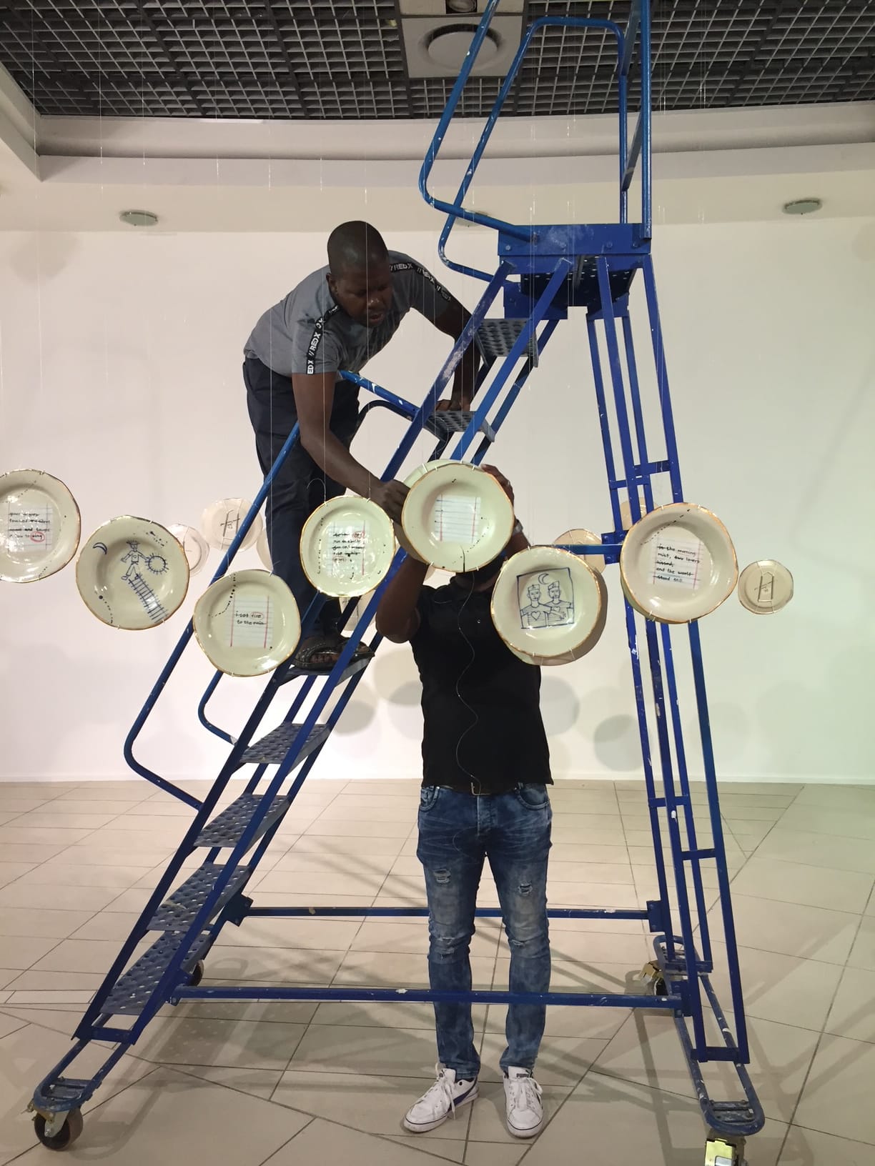 Two men work together hanging decorated plates from nearly invisible strings. One man stands on a blue rolling staircase, reaching out, while the other stands below him. The scene takes place in a modern, white-tiled gallery space.