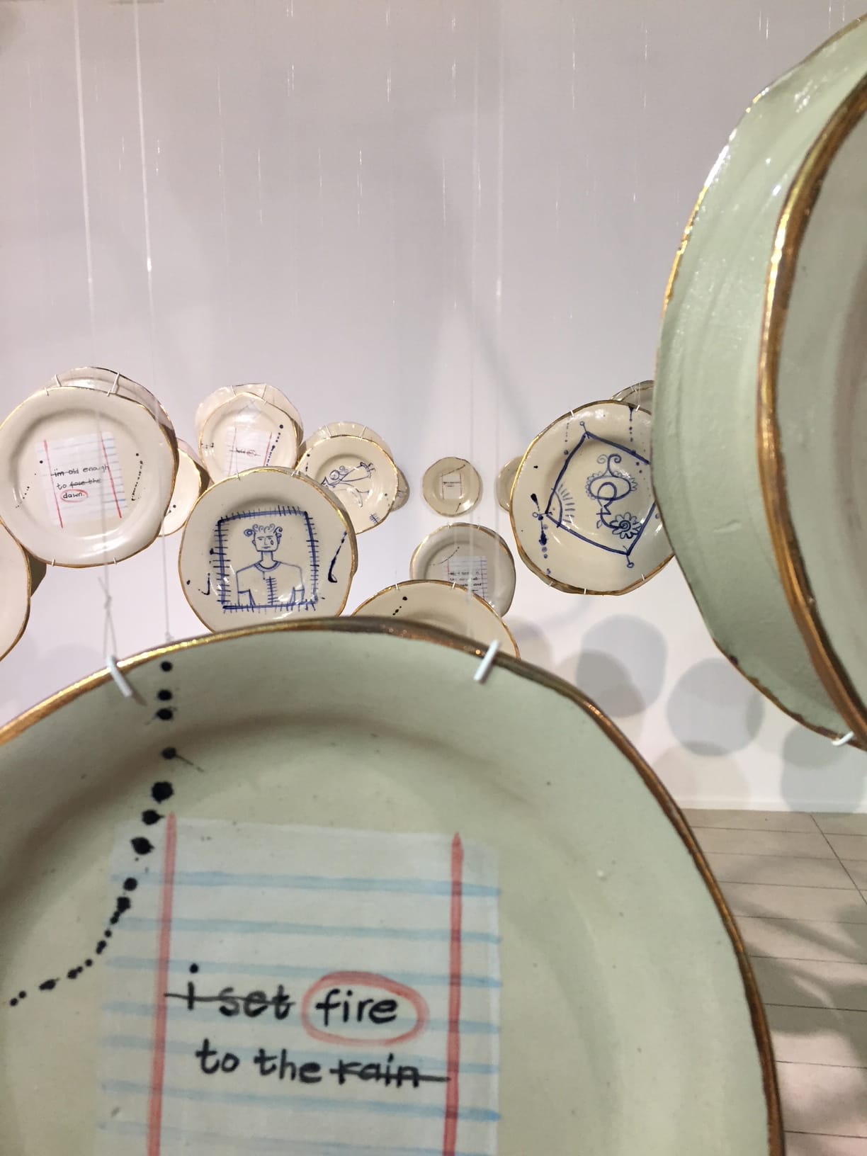 Several ceramic plates hang in rows. The foreground plate displays handwritten text, “I set fire to the rain,” with “fire” circled in red. Other plates feature blue hand-drawn figures, sketches, and lines, all suspended against a white wall.