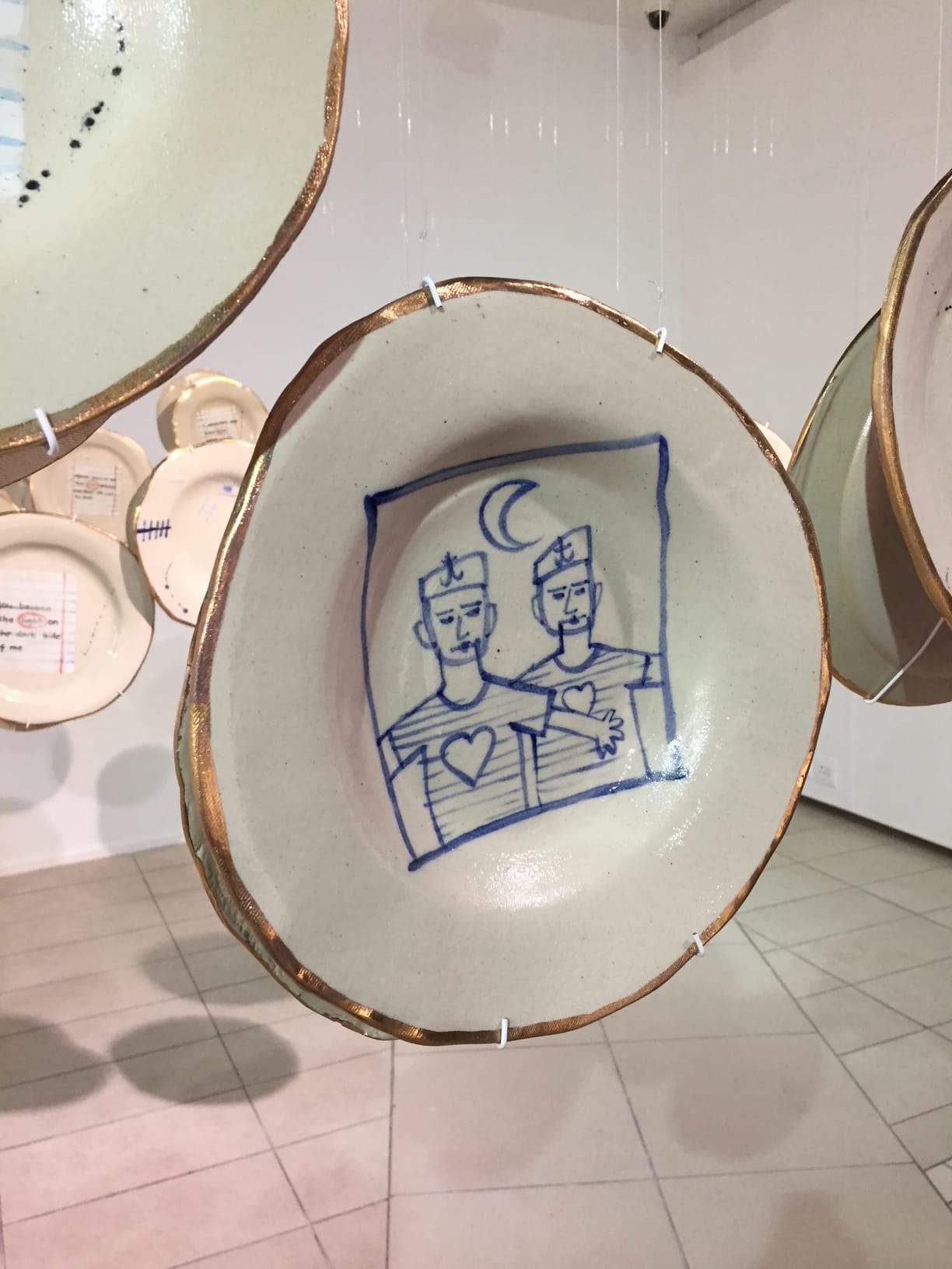 A ceramic plate hangs in an art gallery, featuring a blue line drawing of two people with hearts on their chests and a crescent moon above. The plate's rim is accented with gold, and other decorated plates are visible in the background.