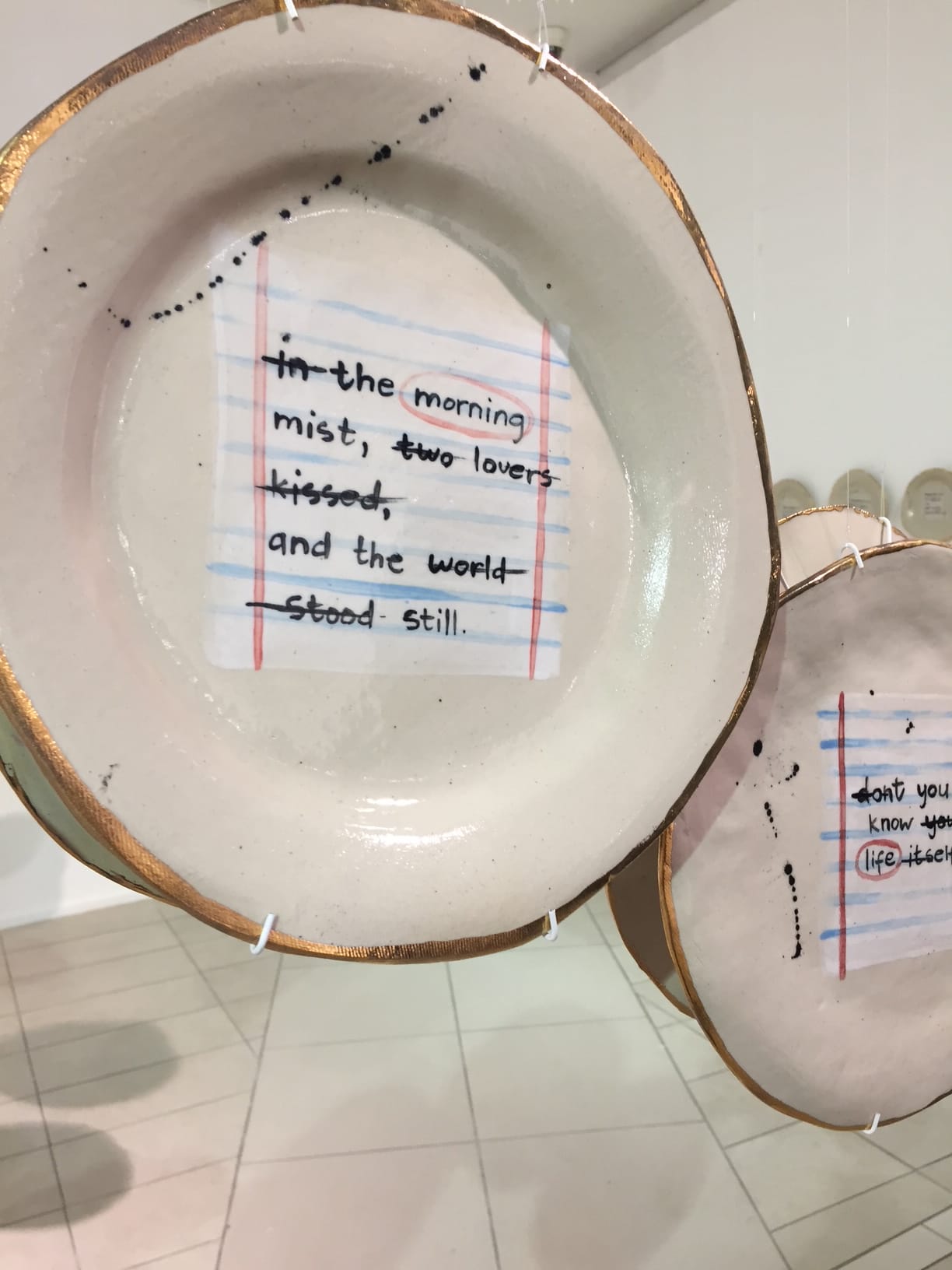 A ceramic plate hangs by strings, decorated to resemble lined exercise book paper. On it is handwritten text: 