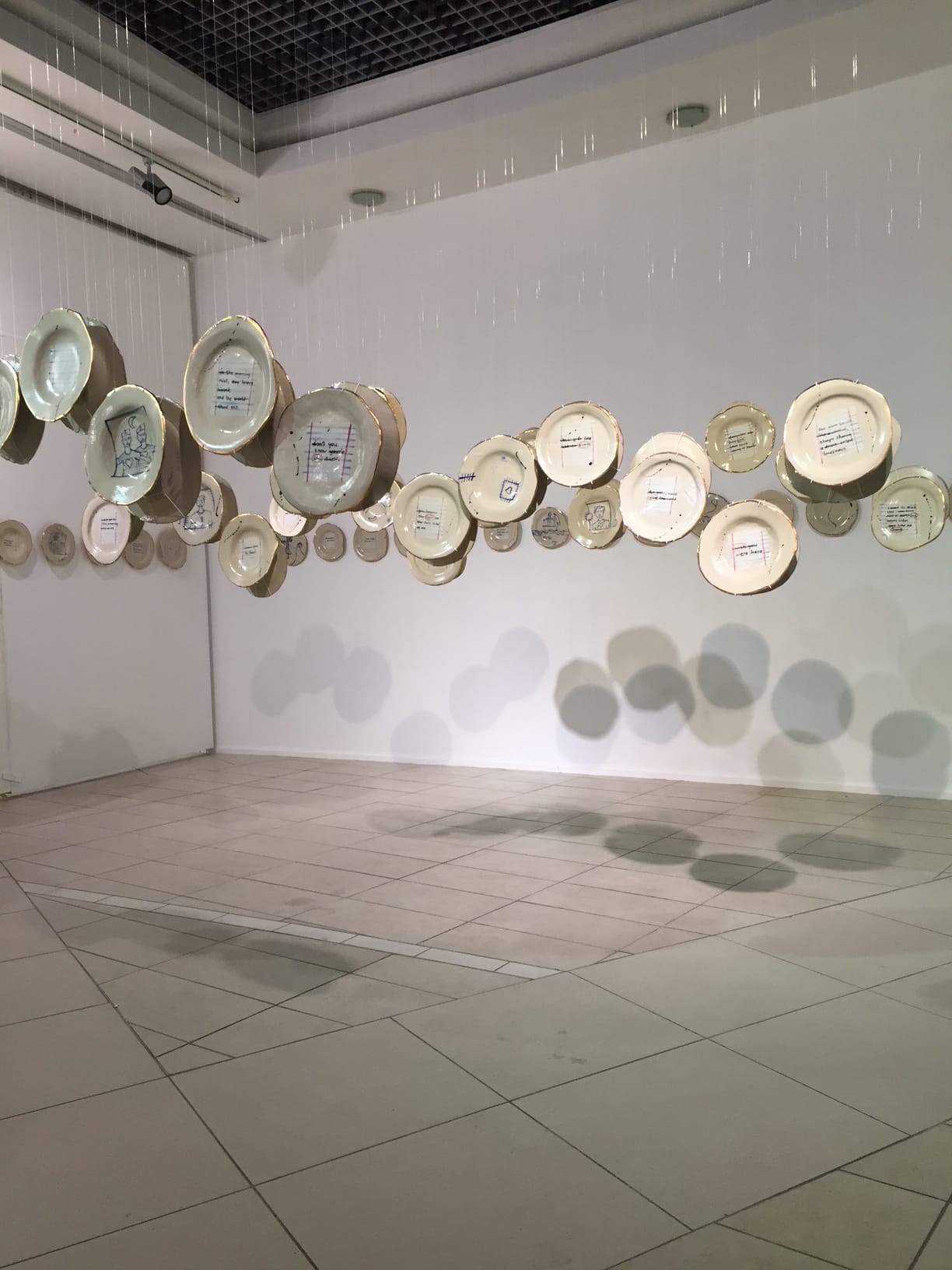 Several cream-coloured ceramic plates are suspended in mid-air by thin, nearly invisible strings in a gallery space. Some plates have drawings or writing on them. Shadows cast by the plates create patterns on the tiled floor and white walls.